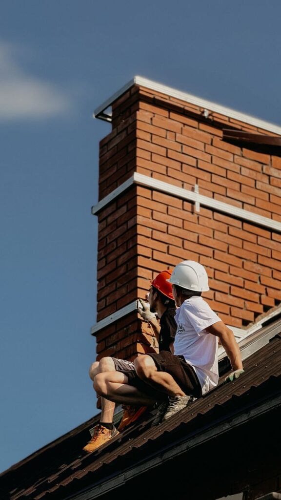 Two construction workers with hard hats repairing a chimney on a house roof under a clear sky.