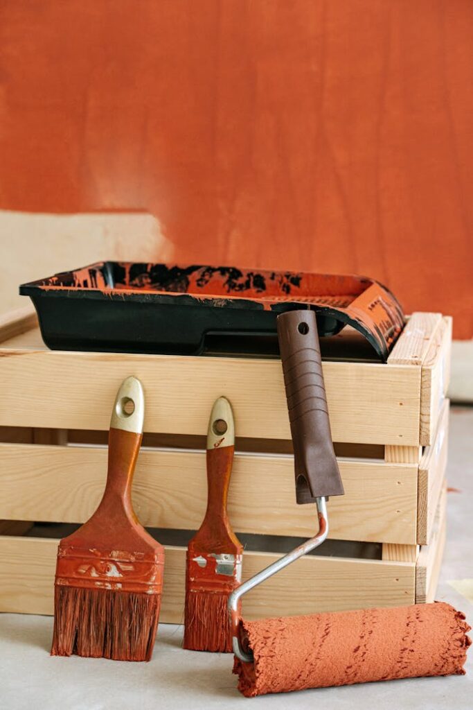 A variety of painting tools including brushes and roller on a wooden crate against a freshly painted wall.